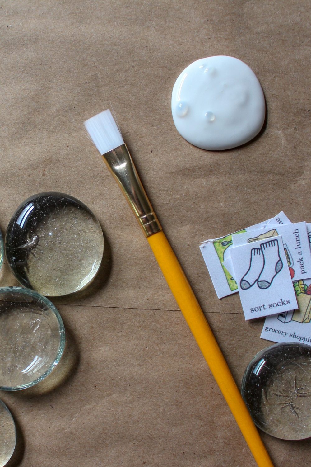 items needed to create chore magnets for kids - pictured school glue, paint brush, clean glass pieces and tasks on paper